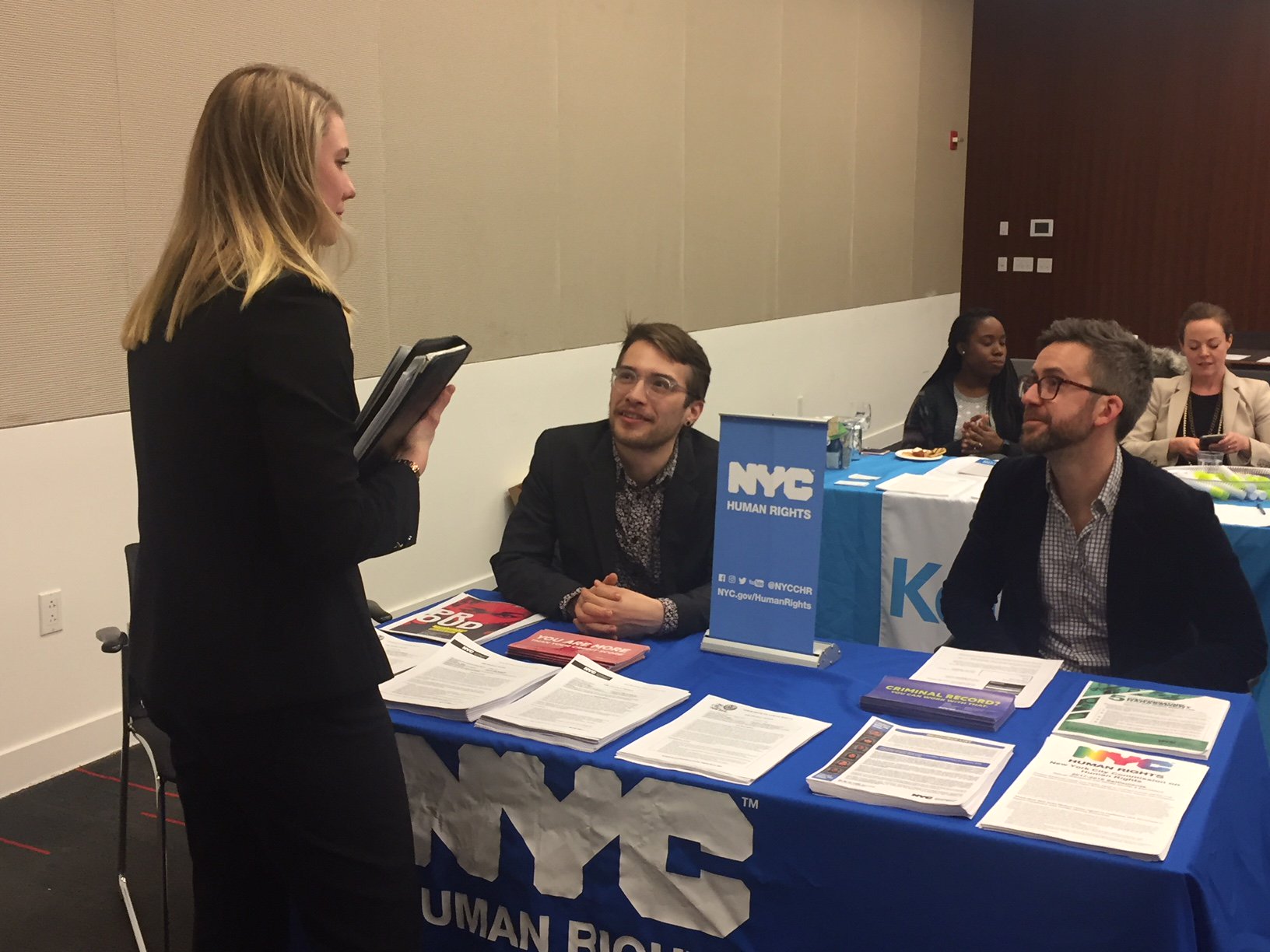 Two Commission staff members sitting at a table speaking with a woman. Tablecloth says, “NYC Human Rights” and there are Commission materials on the table.