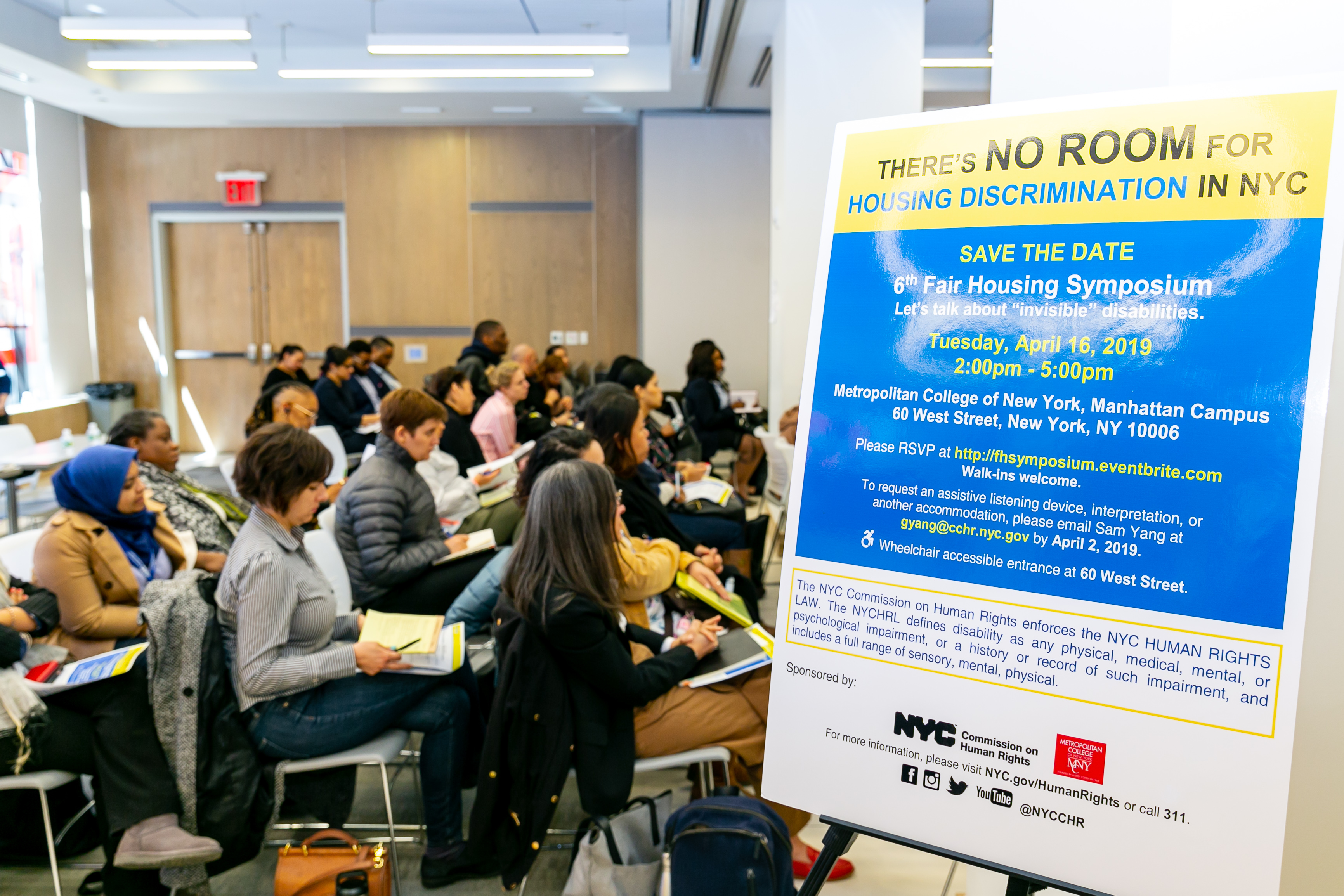 Profile of audience sitting at event next to a sign that reads “There’s No Room For Housing Discrimination in NYC,” 6th Fair Housing Symposium, Tuesday, April 16, 2019.