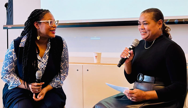 Two women sitting down with microphones in their hands and a screen in the background. One of the women (Commissioner Annabel Palma) is holding the microphone to her mouth and smiling.