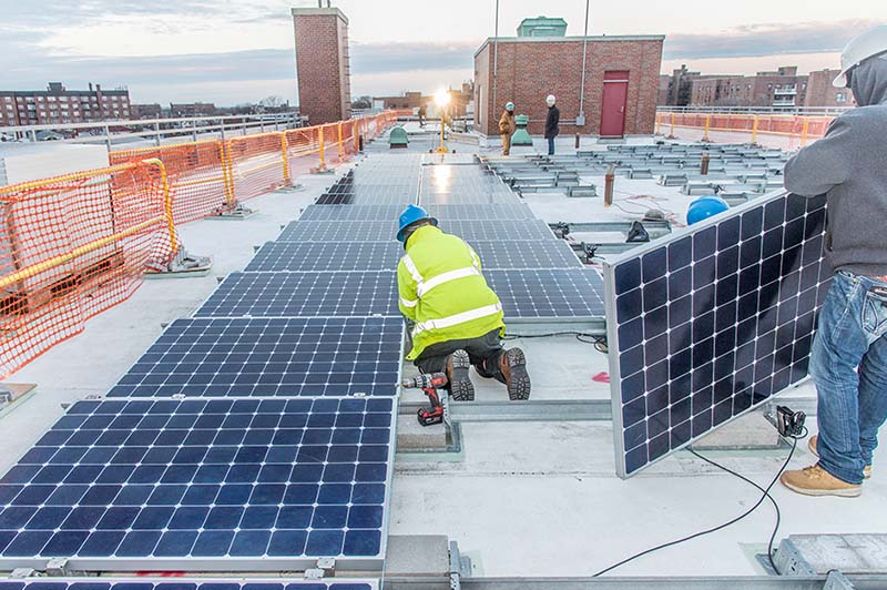 One kneeling worker and one standing worker installing solar panels on a rooftop.