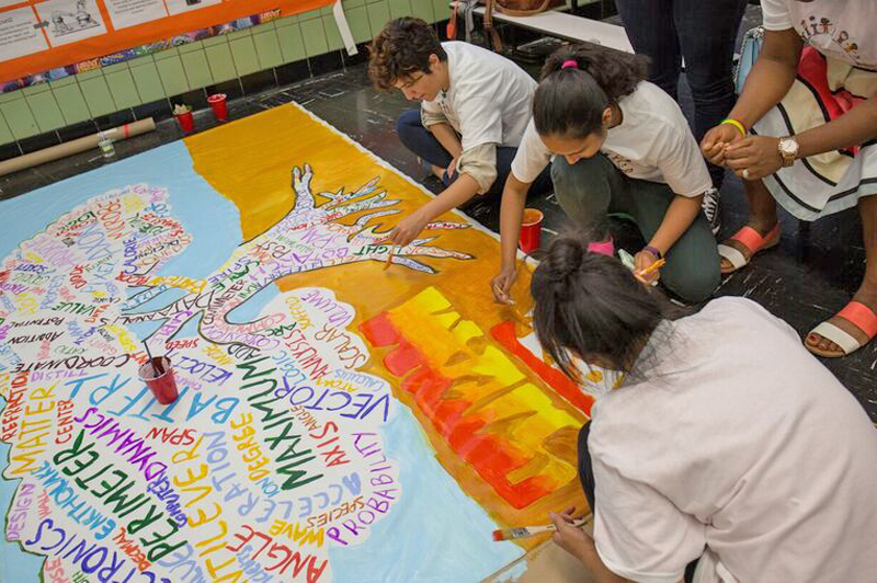 Three people sit on the floor and paint on a large piece of paper for a mural.