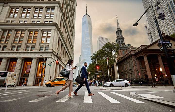Two people crossing a city street in the cross walk.  In the background are a historic building, a modern skyscraper, and a historic church.