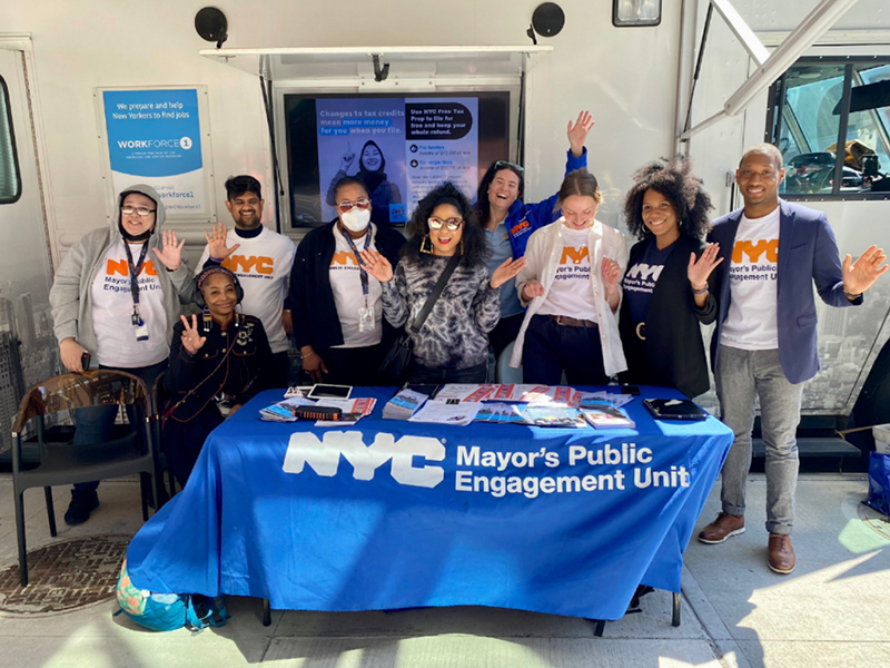 PEU staff wearing t-shirts standing in front of a van and a table that says the Mayor's Public Engagement Unit. In the middle is Angela Yee