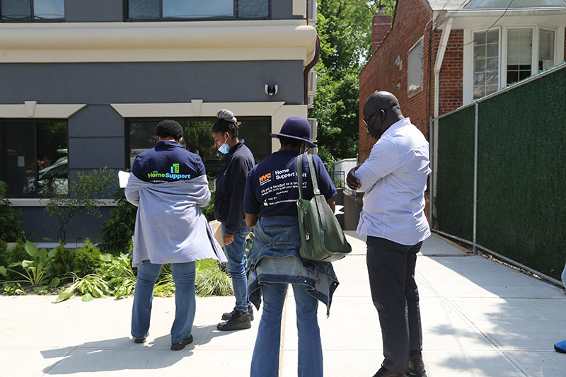 Two Home Support Unit Specialists talk with people outside of an apartment building. Their backs are turned to the camera and the backs of their shirts have information about the Home Support Unit