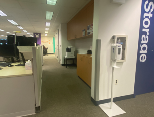 There is a pathway. On the left is a cubicle with a computer and on the right are cabinets and a printer. On the left there is a door with the word Storage painted in white letters.
