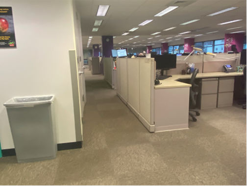 There is a pathway. On the right is a cubicle with a computer are doors. There are also more cubicles with purple and pink columns in the background.
