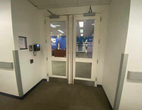 MOCS' main entrance that has two doors with glass windows. One of the glass windows has the NYC emblem and the Mayor's Office of Contract Services in blue letters. There is a phone and intercom on the left of the doors.