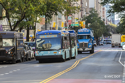 A blue M14 Select Bus Service bus travels along a traffic-free 14th Street followed by a blue truck.