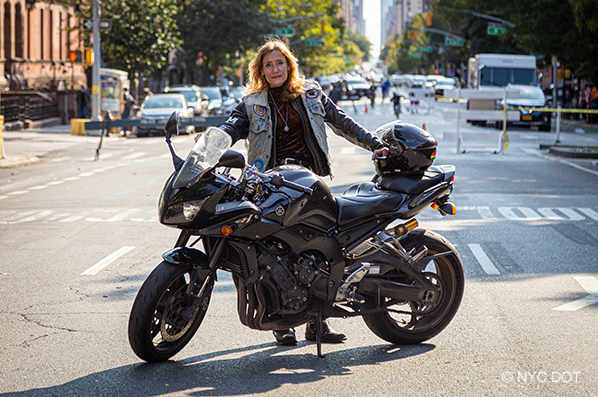 A woman wearing motorcycle gear stands beside a motorcycle parked on a street closed to traffic. Her helmet is resting on the motorcycles seat.