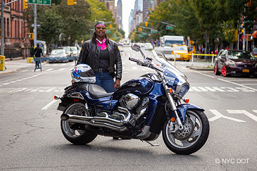 A woman wearing motorcycle gear, stands behind a motorcycle parked on a street closed to traffic. Her helmet is resting on the seat.