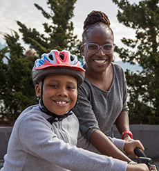 A woman and a child smile at the camera. The child is wearing a helmet and the woman is helping him ride a bike.