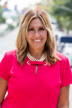 Portrait of NYC DOT’s Staten Island Borough Commissioner, Roseann Caruana standing outside on a sunny day, wearing a magenta dress with pearls