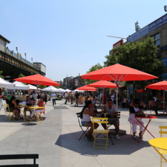 People sit on chairs around tables with red umbrellas at Corona Plaza. 