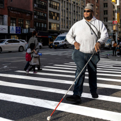 A man using a white and red cane crosses a marked crosswalk.