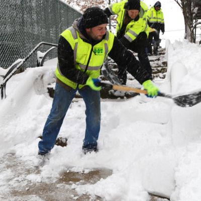 Three people wearing fluorescent jackets shovel snow from a step street.