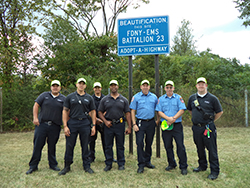Seven Fire Department/Emergency Medical Service Members standing in front of their Adopt a Highway Beautification sign on the side of the West Shore Expressway. The sign reads  FDNY – EMS Battalion 23