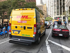 An orange DHL van with a sticker promoting the vehicle as '“100% Electric”' drives along a NYC street.
