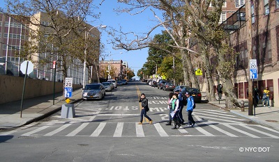 A group of students walk in a crosswalk, across a two lane road with two white “Speed Limit 20” signs and two blue “Neighborhood Slow Zone” signs on either side of the road.