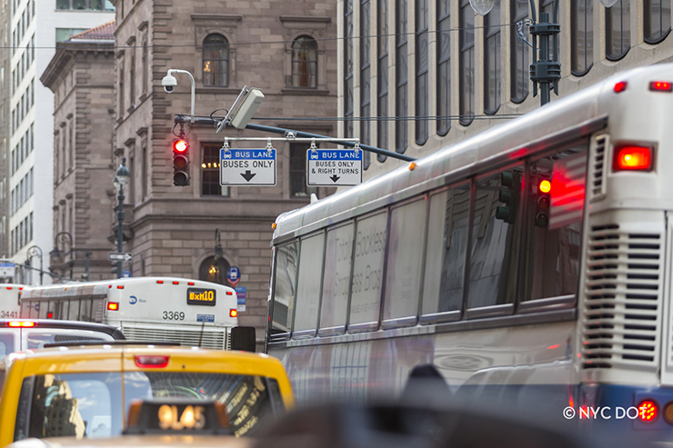 traffic waits at a red traffic signal on a busy midtown street. Buses are lined up in a lane, under “Bus Lane: Buses Only” signs hanging from a traffic signal’s pole.