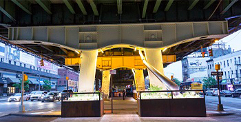 Below an elevated roadway, bright paint, lights and large, illuminated planters brighten the area near a sidewalk.