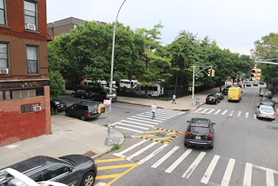 A car turns left around a slow turn wedge treatment, helping the driver to see a pedestrian walking in the crosswalk. Location is Lexington Ave & Marcy Ave in Brooklyn, NY