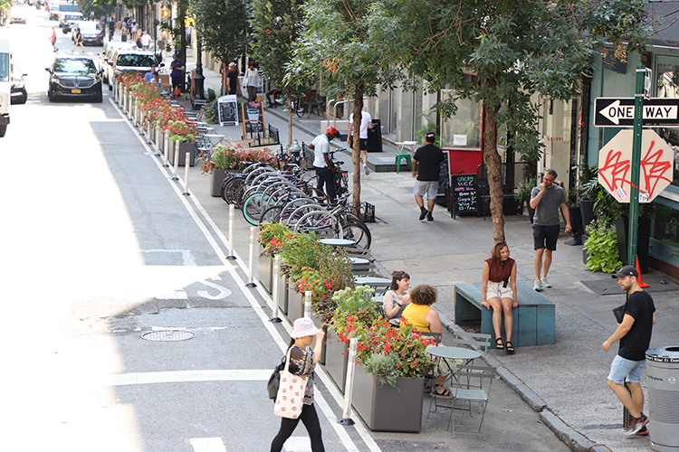 Planters and flexible posts line a long extended sidewalk area in a roadway. The space is filled with bike parking and tables and chairs. Two people sit at one of the tables on a warm day in NYC.