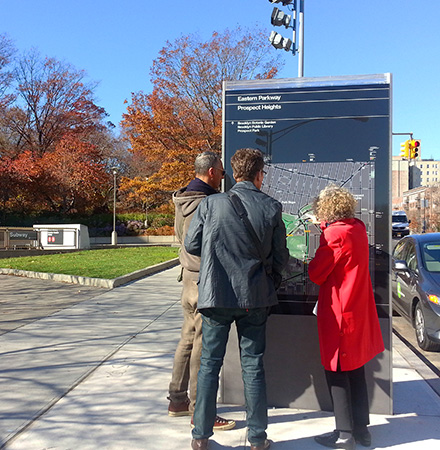 Outside a park on a wide sidewalk, a group of people look at a tall, wide sign with a neighborhood map