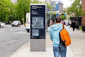 A man carrying an orange backpack walks on a wide sidewalk, past a tall sign with two maps on it