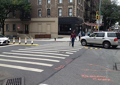 A car turns left around a basic hardened centerline treatment, helping the driver to see a pedestrian walking in the crosswalk. Location is West End Ave & 96 St in Manhattan, NY.