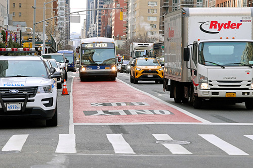 An MTA bus travels along a red 'Bus Only' lane on a wide avenue in Manhattan